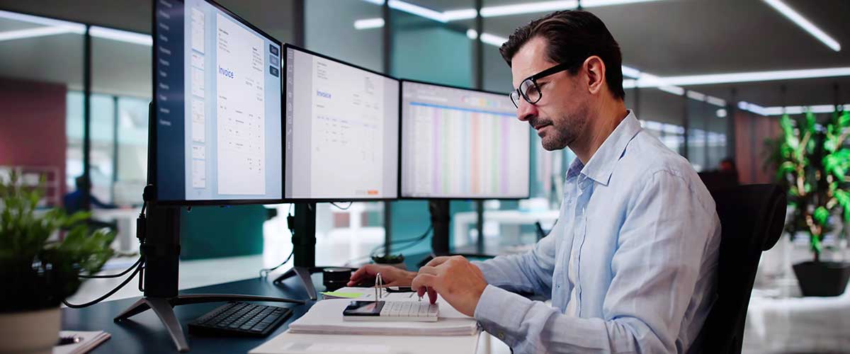Man using calculator at desk with three monitors.