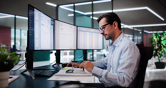 Man using a calculator at desk with three monitors.